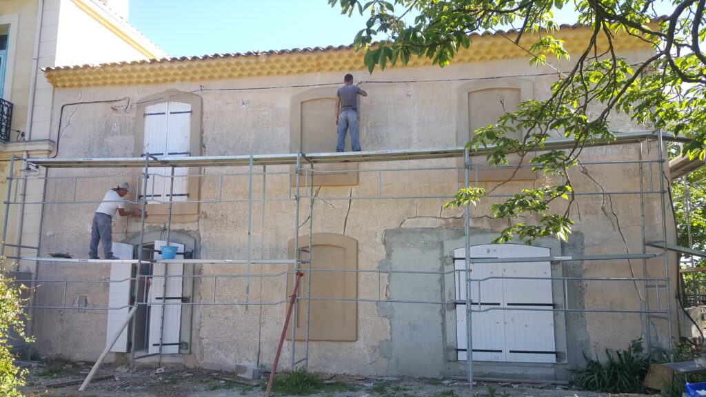 Image après rénovation : Façade rénovée avec nouvel enduit ocre, fenêtres protégées par film plastique, échafaudage en place, montrant un chantier de rénovation en cours avec finitions professionnelles.