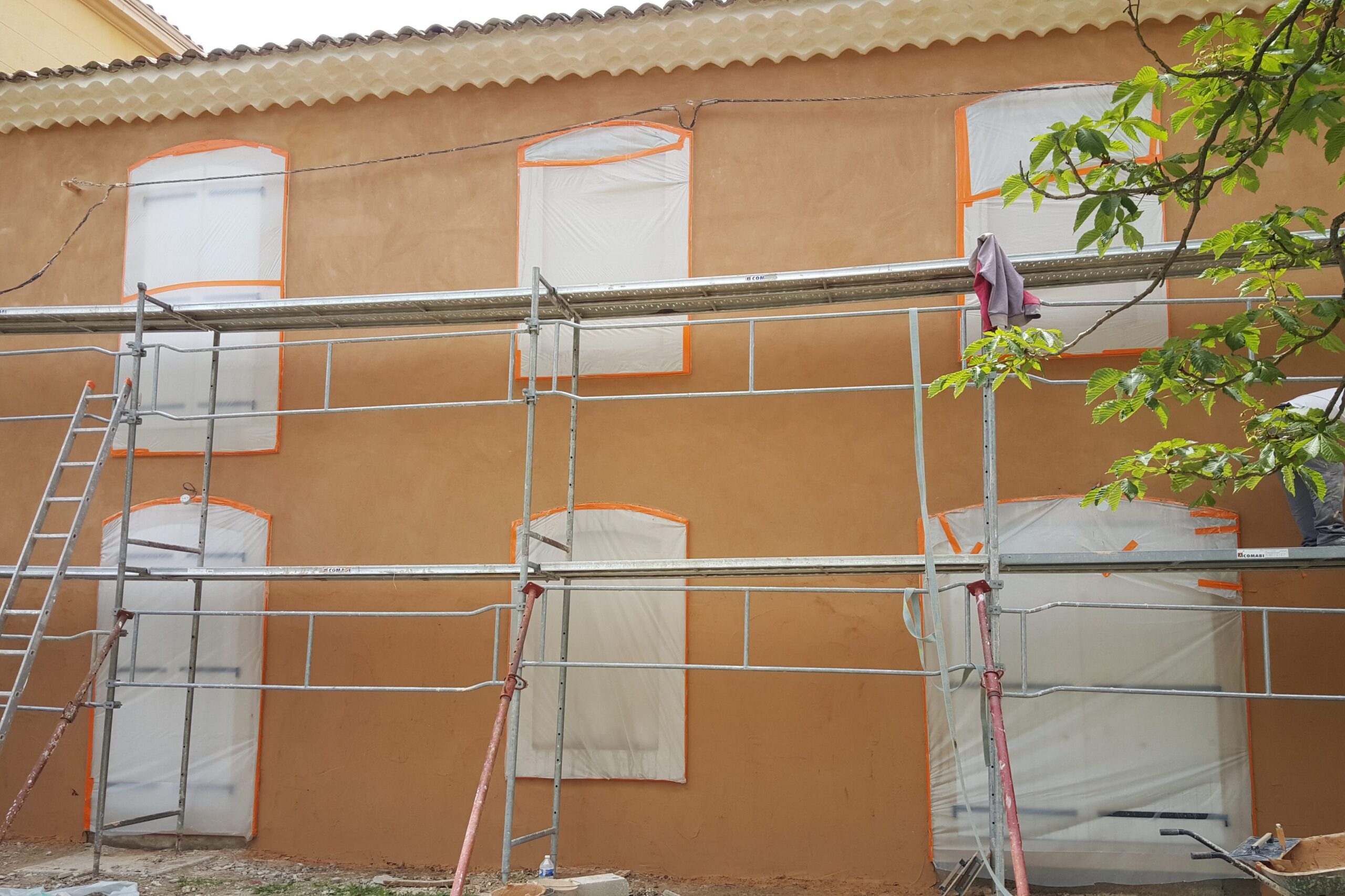 Image après rénovation : Façade rénovée avec nouvel enduit ocre, fenêtres protégées par film plastique, échafaudage en place, montrant un chantier de rénovation en cours avec finitions professionnelles.
